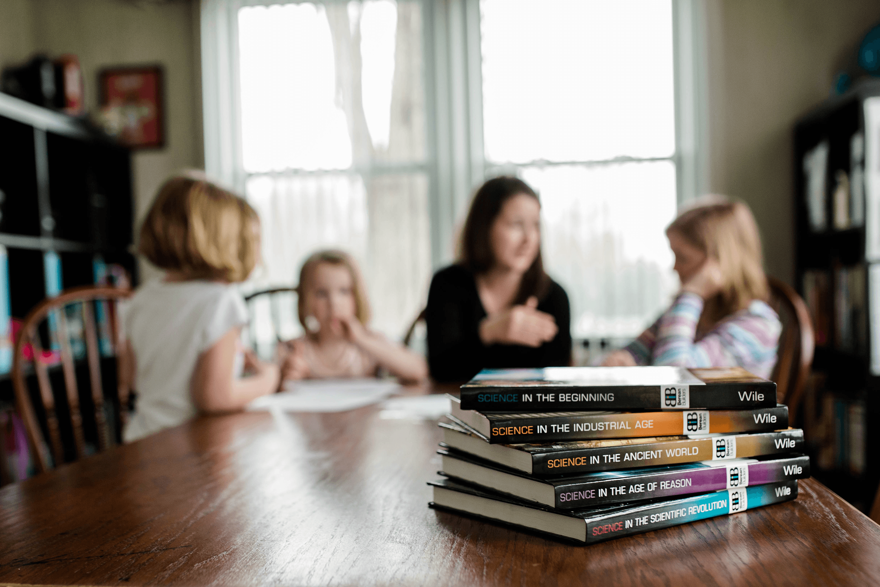 A stack of Berean Builders Elementary courses sits atop a dining table in the foreground while a mother speaks with her daughters around the table in the background